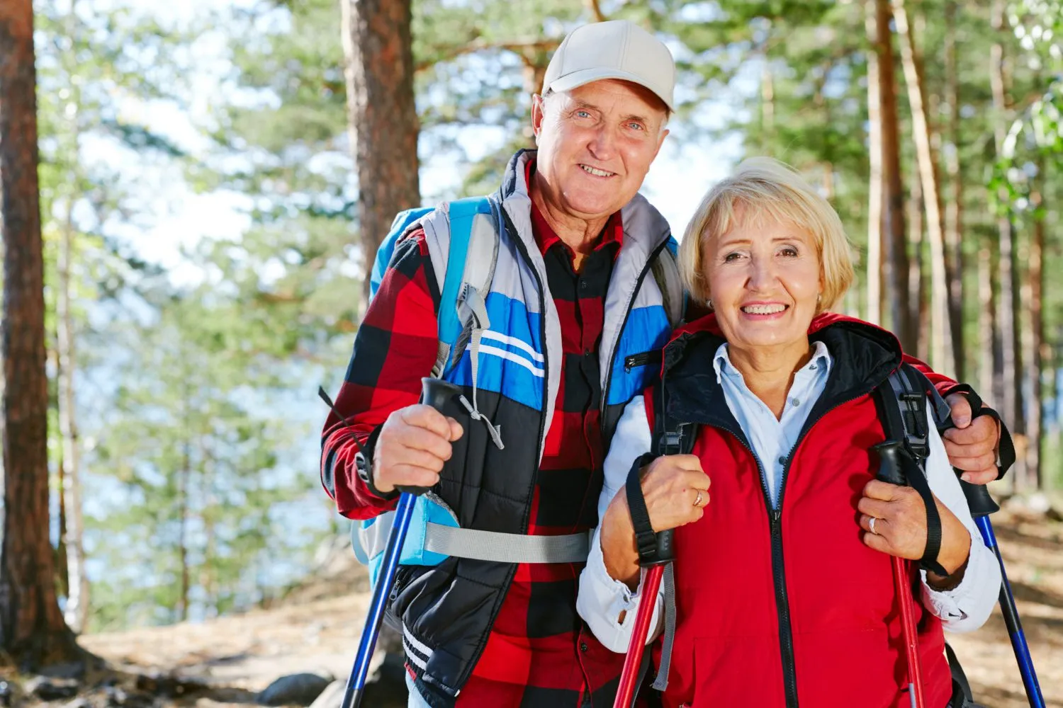 Two hikers in colorful jackets stand together in a sunlit forest, holding trekking poles, surrounded by tall trees Two hikers in colorful jackets stand together in a sunlit forest, holding trekking poles, surrounded by tall trees